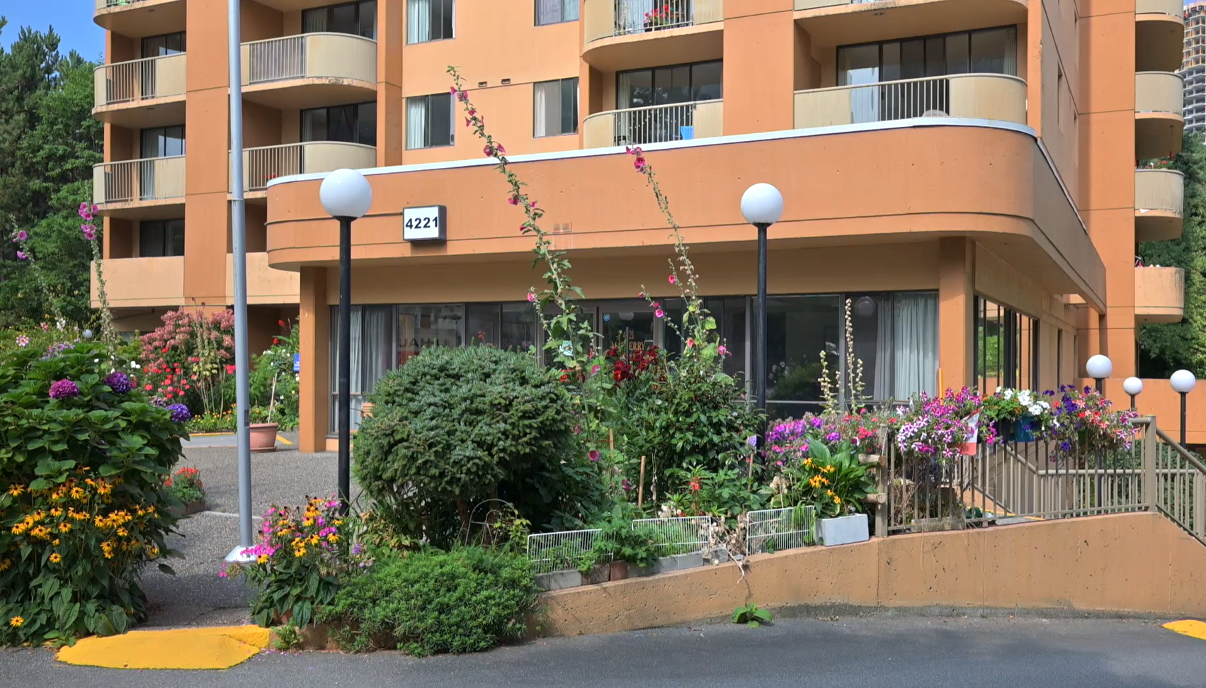 Front entrance of Post 83 Housing Co-op in Burnaby, featuring landscaped gardens with colorful flowers and shrubs in front of a peach-colored multi-level building.