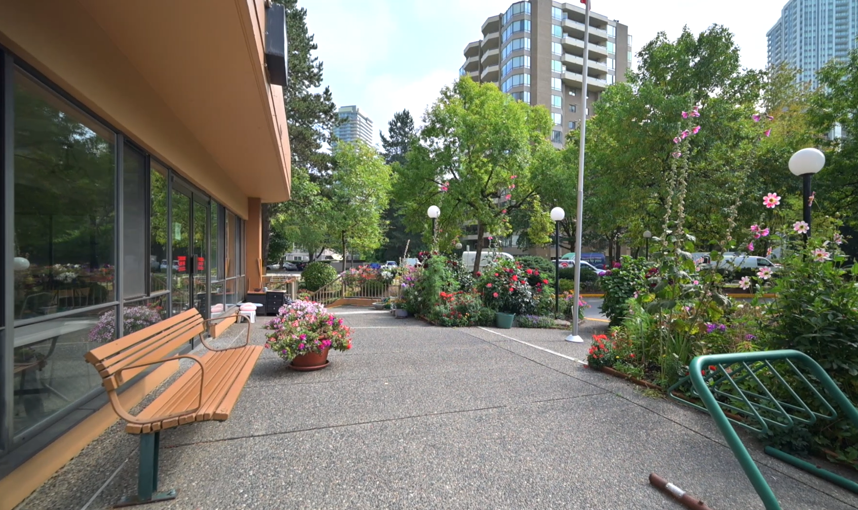 Outdoor seating area at Post 83 Housing Co-op in Burnaby, featuring a wooden bench, potted flowers, and landscaped greenery with high-rise buildings in the background.