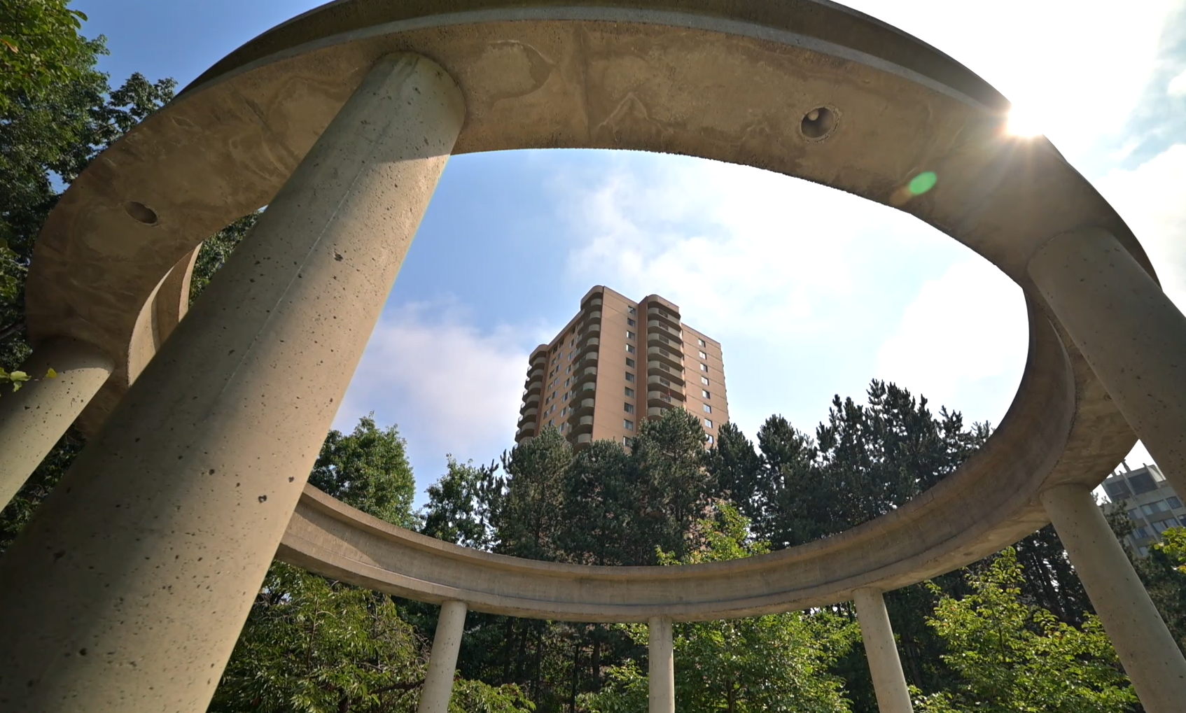Upward view through a circular concrete structure at Post 83 Housing Co-op in Burnaby, with a high-rise building framed against a bright sky and surrounding trees.