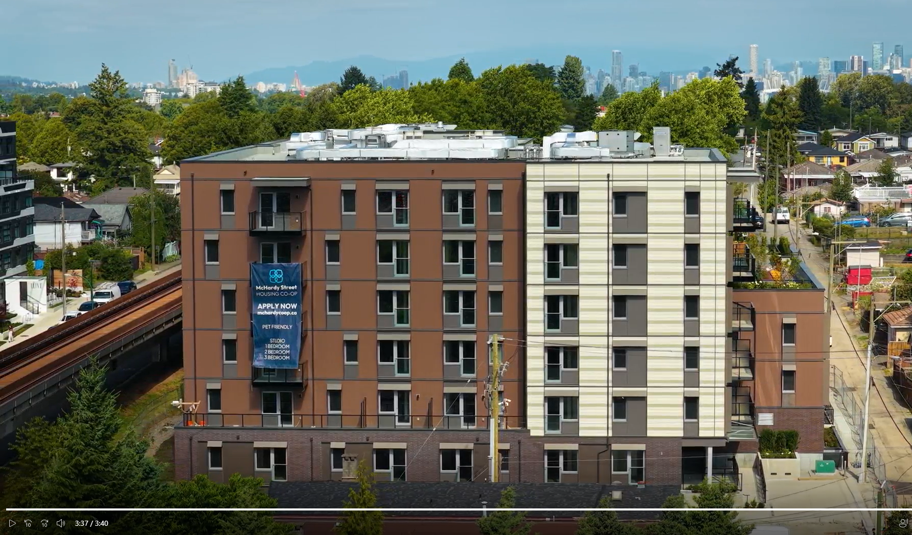 Six-story apartment building with brown and beige panels, banner reading ‘Apply Now,’ trees and city skyline in background.